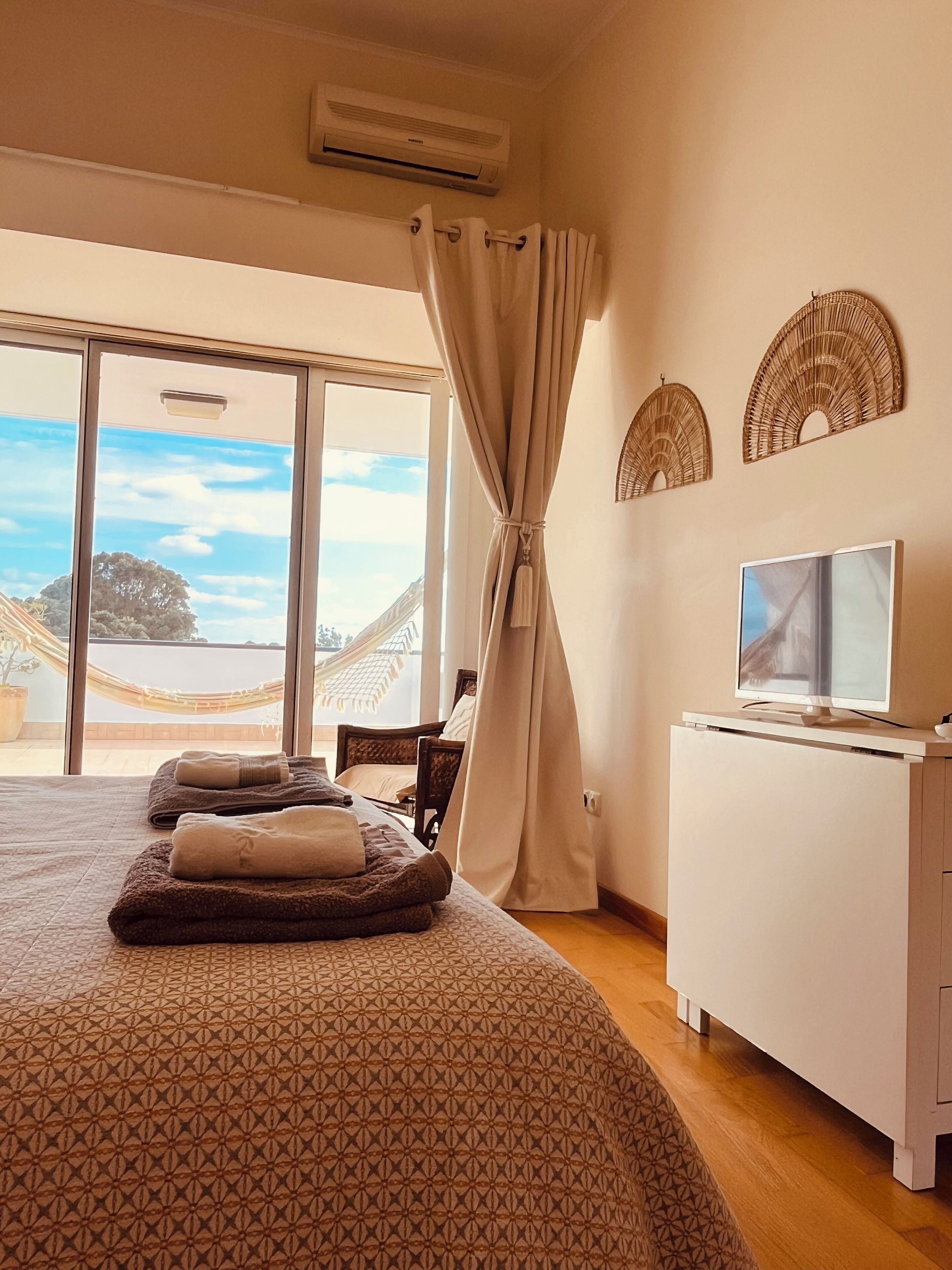 Une chambre avec un grand lit, un téléviseur posé sur une commode blanche et une vue sur l’extérieur grâce à une grande fenêtre.
