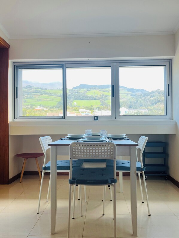 A dining area with a table set for two, two chairs, and a view of green hills through large windows.