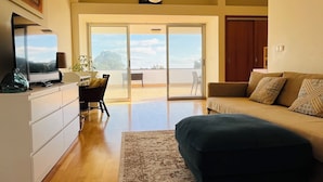 A spacious living room with a large bed, a desk, and a view through a sliding glass door to an outdoor area.
