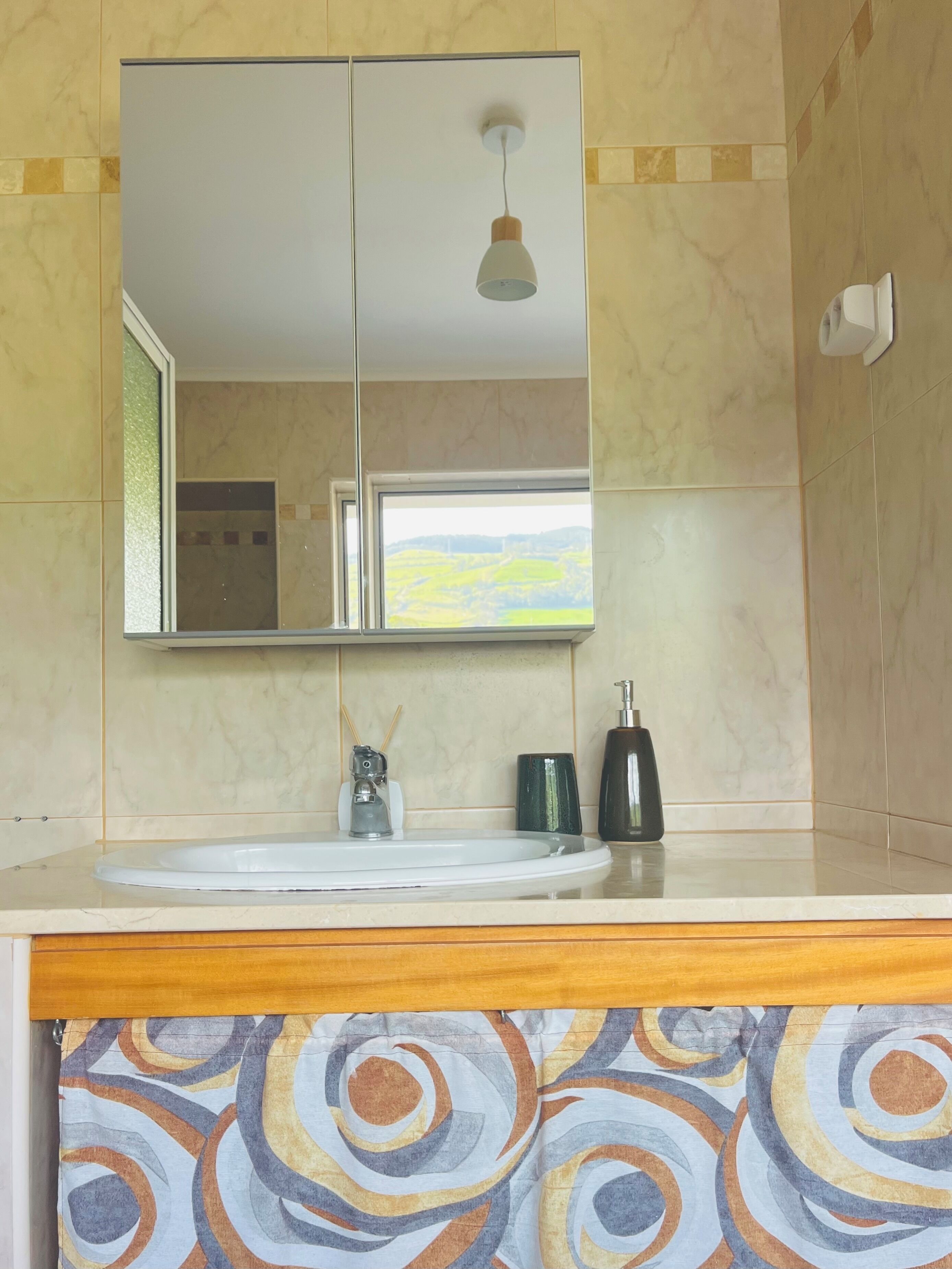 Een badkamer met spiegel, wastafel en een raam met uitzicht op groene velden.