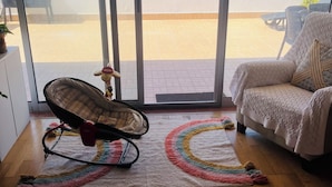 A room with a view through a sliding glass door to a balcony, a rocking chair, a white sofa, and a colorful rug.