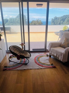 Une pièce avec vue sur un balcon, accessible par une porte-fenêtre coulissante, comprenant un fauteuil à bascule, un canapé blanc et un tapis coloré.