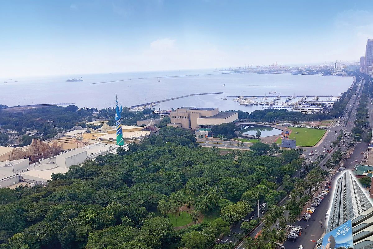 Paisagem costeira urbana com um edifício imponente e uma torre listrada em azul e branco, uma marina com barcos e uma estrada ladeada por palmeiras.