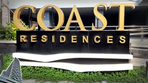 Une enseigne indiquant « COAST RESIDENCES », avec de grandes lettres dorées sur un fond noir, le tout entouré de verdure.