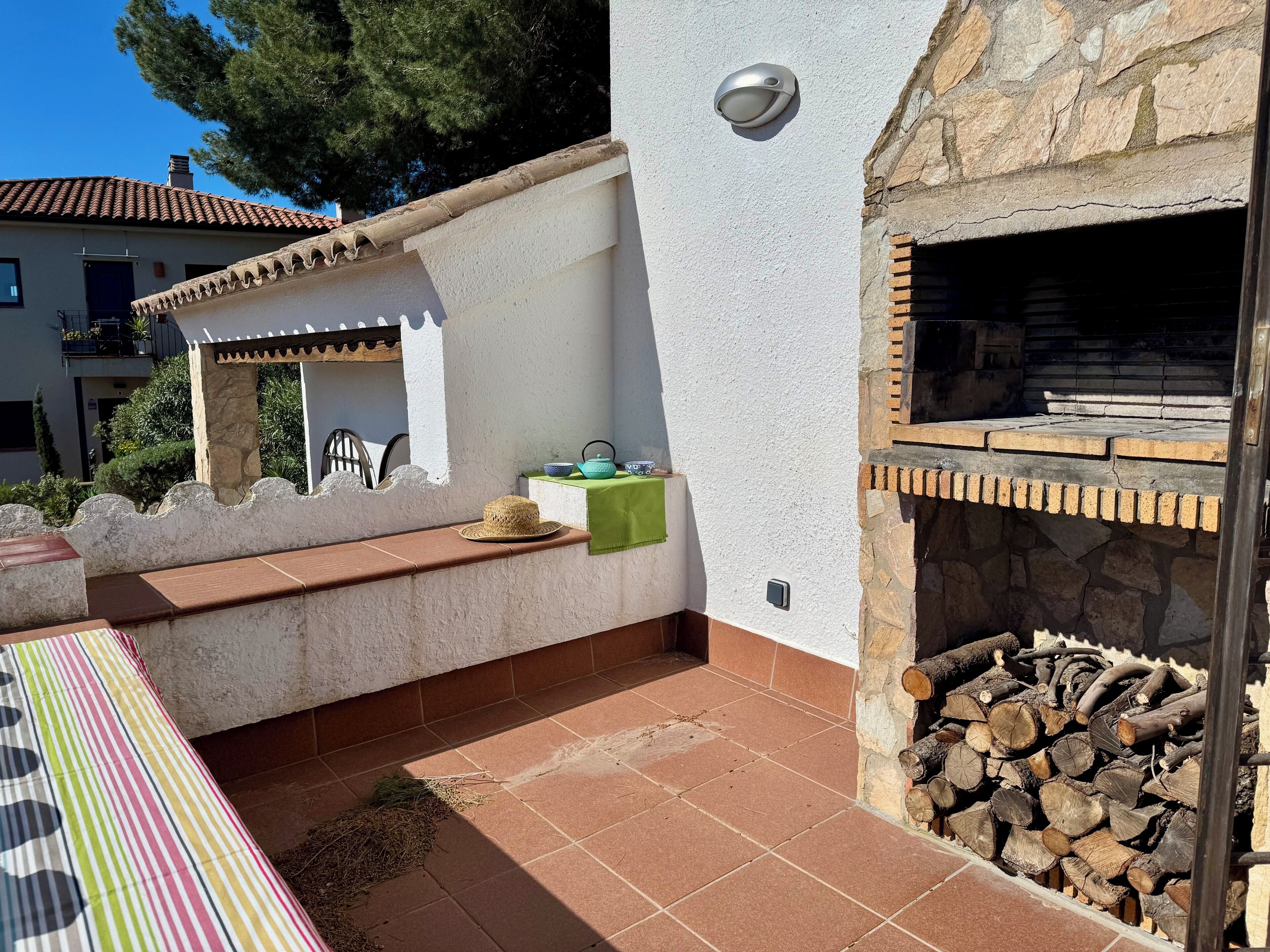 A patio with a tiled floor, a wooden bench, a table with a striped tablecloth, a stone wall, a metal grill, and a stack of firewood.