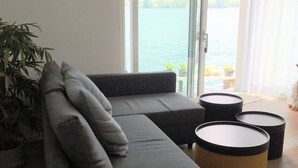 Ein modernes Wohnzimmer mit einer grauen Eckcouch, einer Topfpflanze und Blick auf ein Gewässer durch große Fenster.