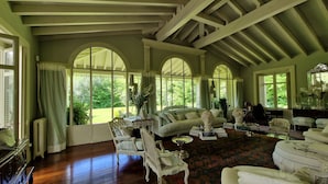 Ein geräumiges Wohnzimmer mit hohen Decken, großen Bogenfenstern und Blick auf den Garten. Es befinden sich mehrere antike Möbelstücke im Raum, darunter ein Sofa, Sessel und ein Tisch.