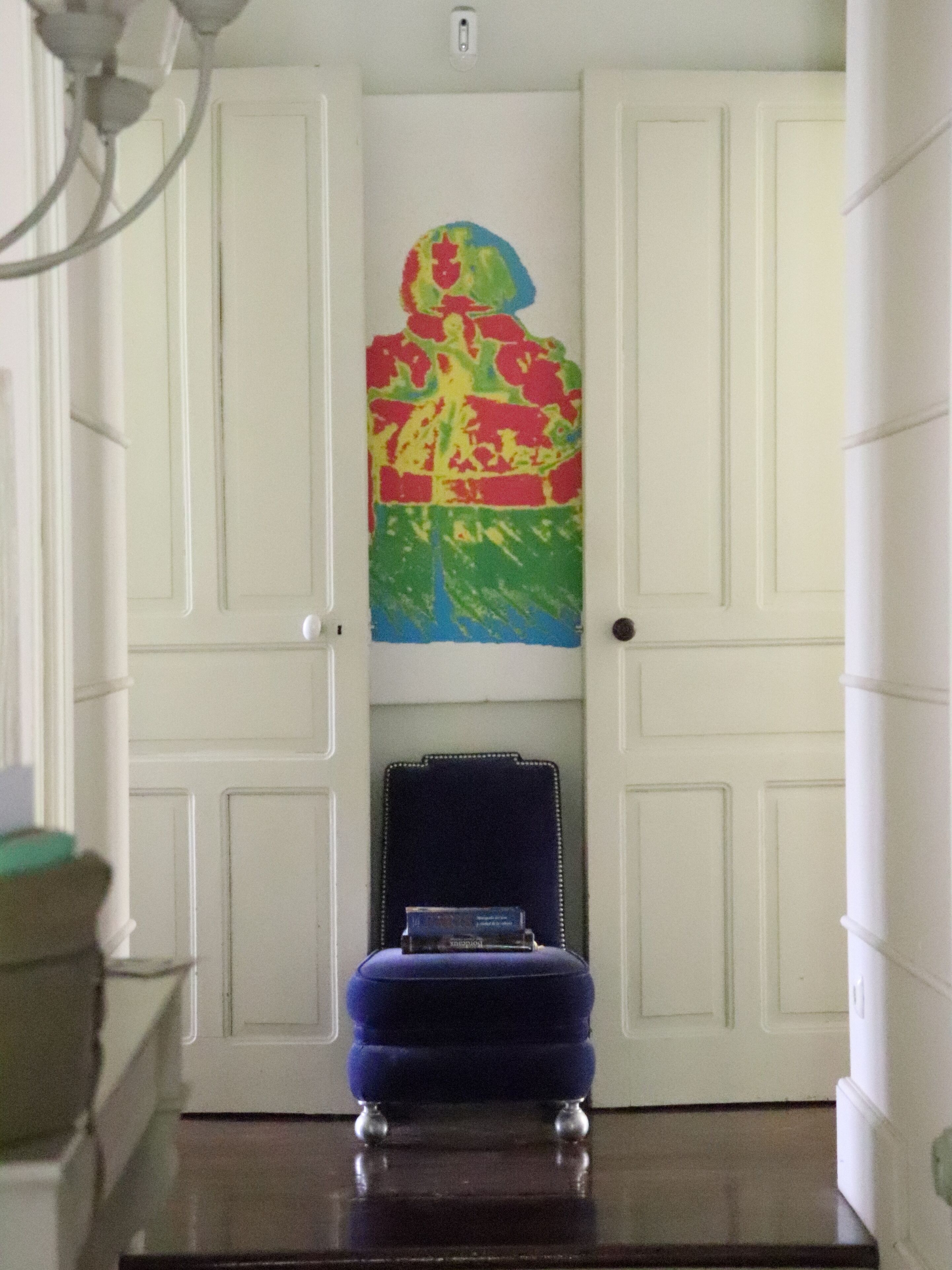 A hallway with a blue ottoman, a colorful abstract painting on the wall, and a black suitcase on a shelf.