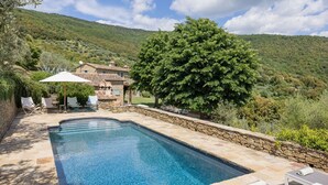 Zona de piscina con muros de piedra, sillones reclinables y un árbol grande.
