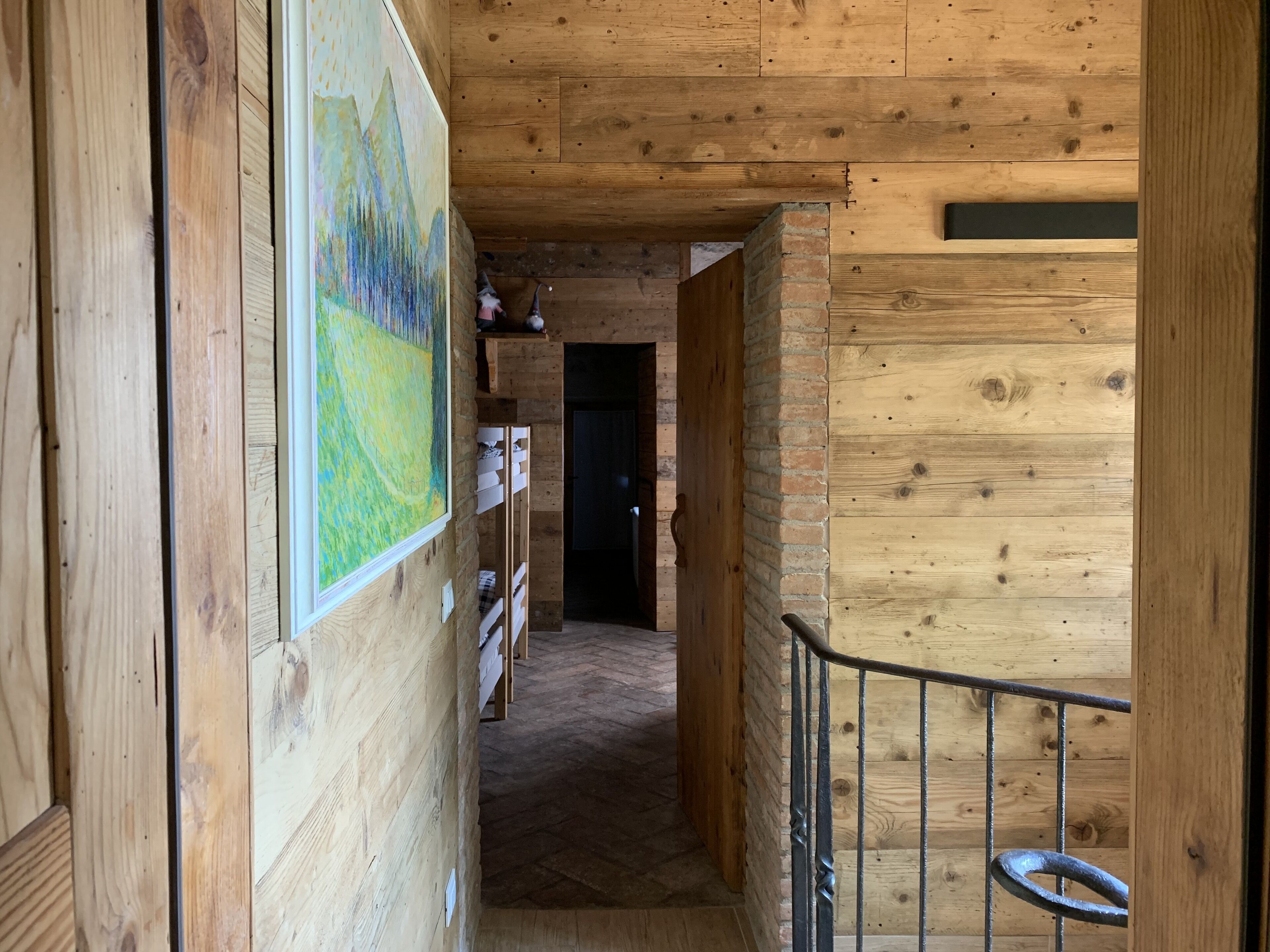 A wooden interior hallway with a painting on the left, a brick wall on the right, and a metal railing in the foreground.