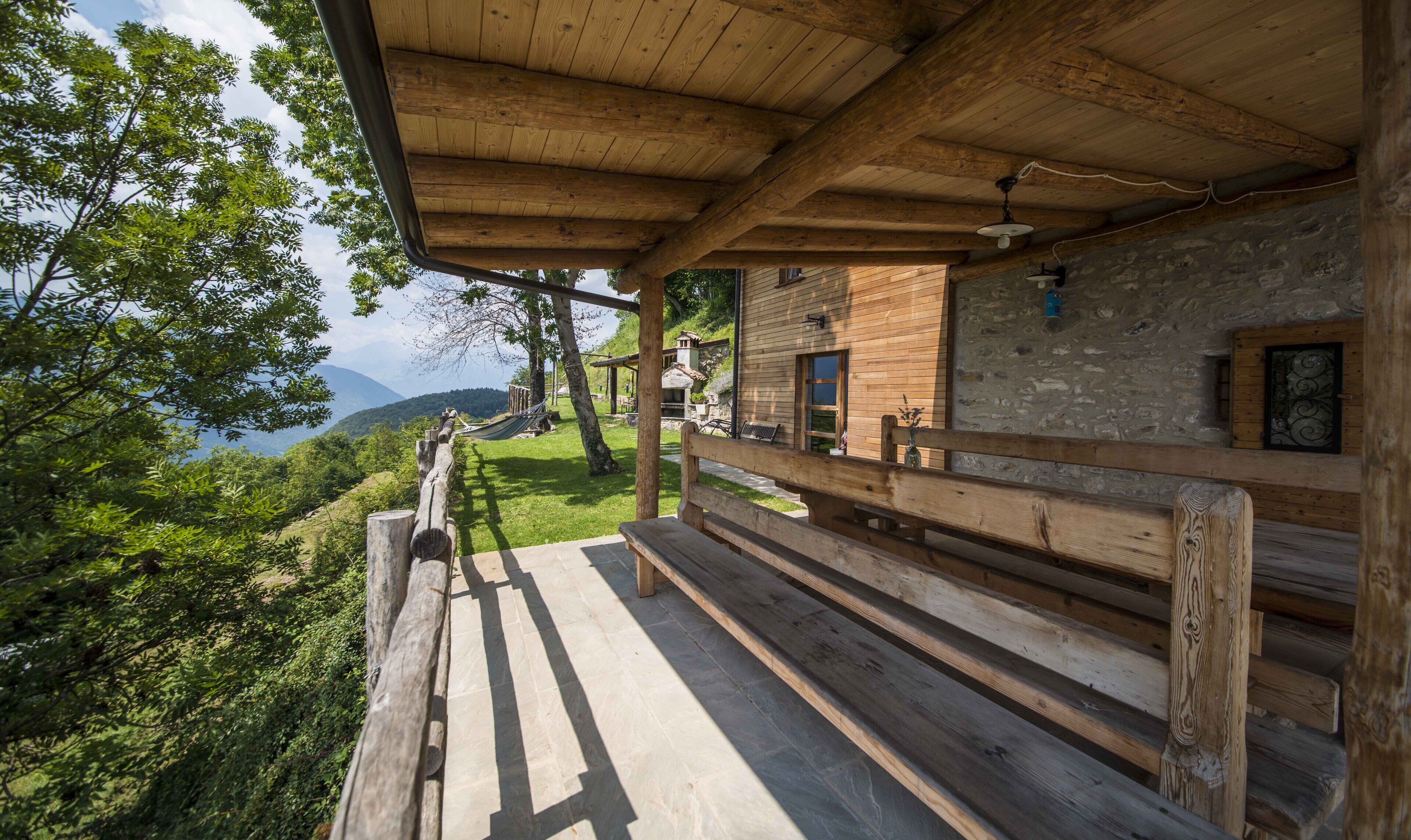 Una baita di legno con parete in pietra e terrazza in legno che si affaccia su un paesaggio montuoso.