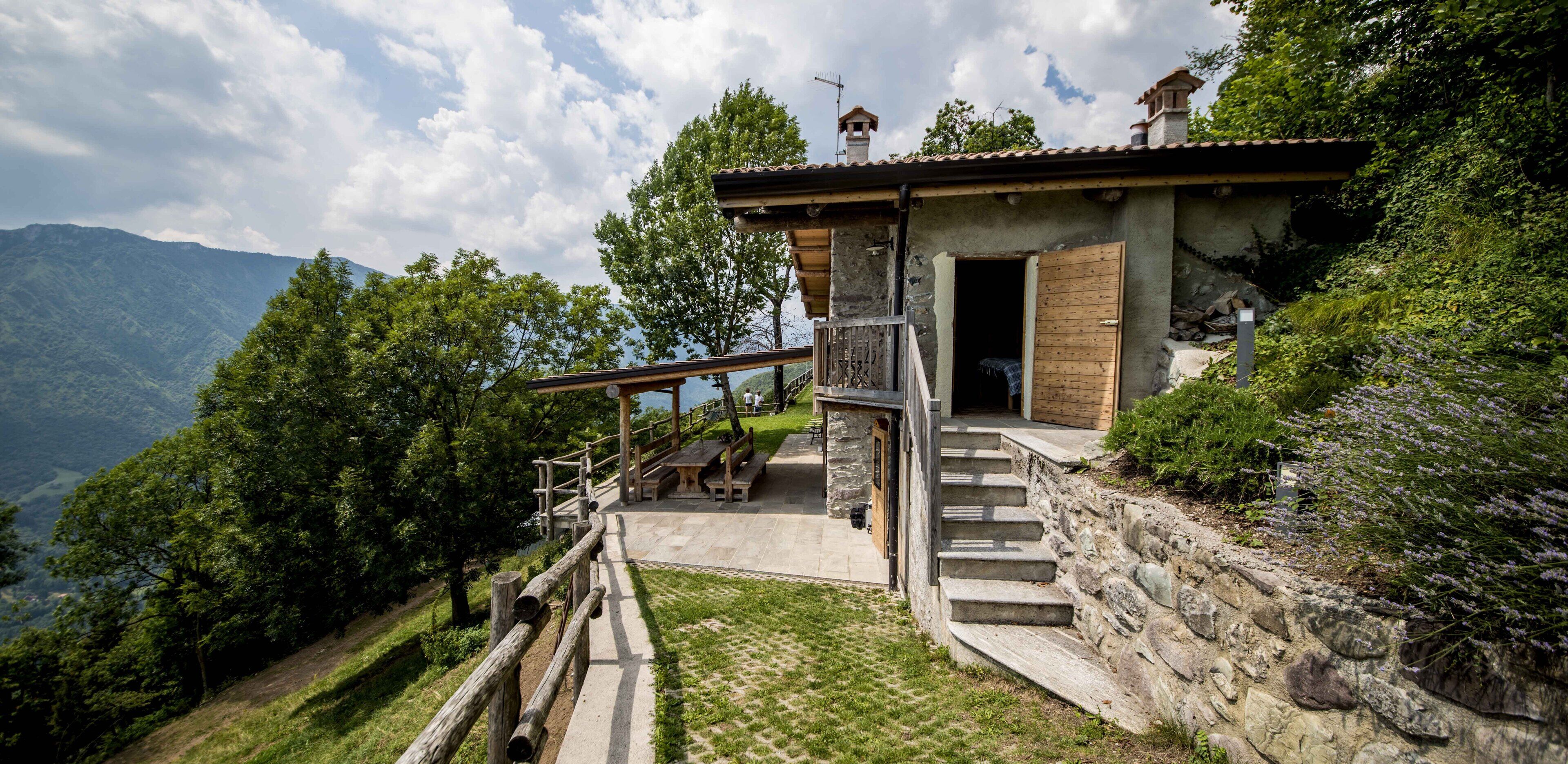 Uma cabana rústica na montanha, com degraus de pedra que levam a uma porta de madeira, cercada por vegetação exuberante e montanhas ao fundo.