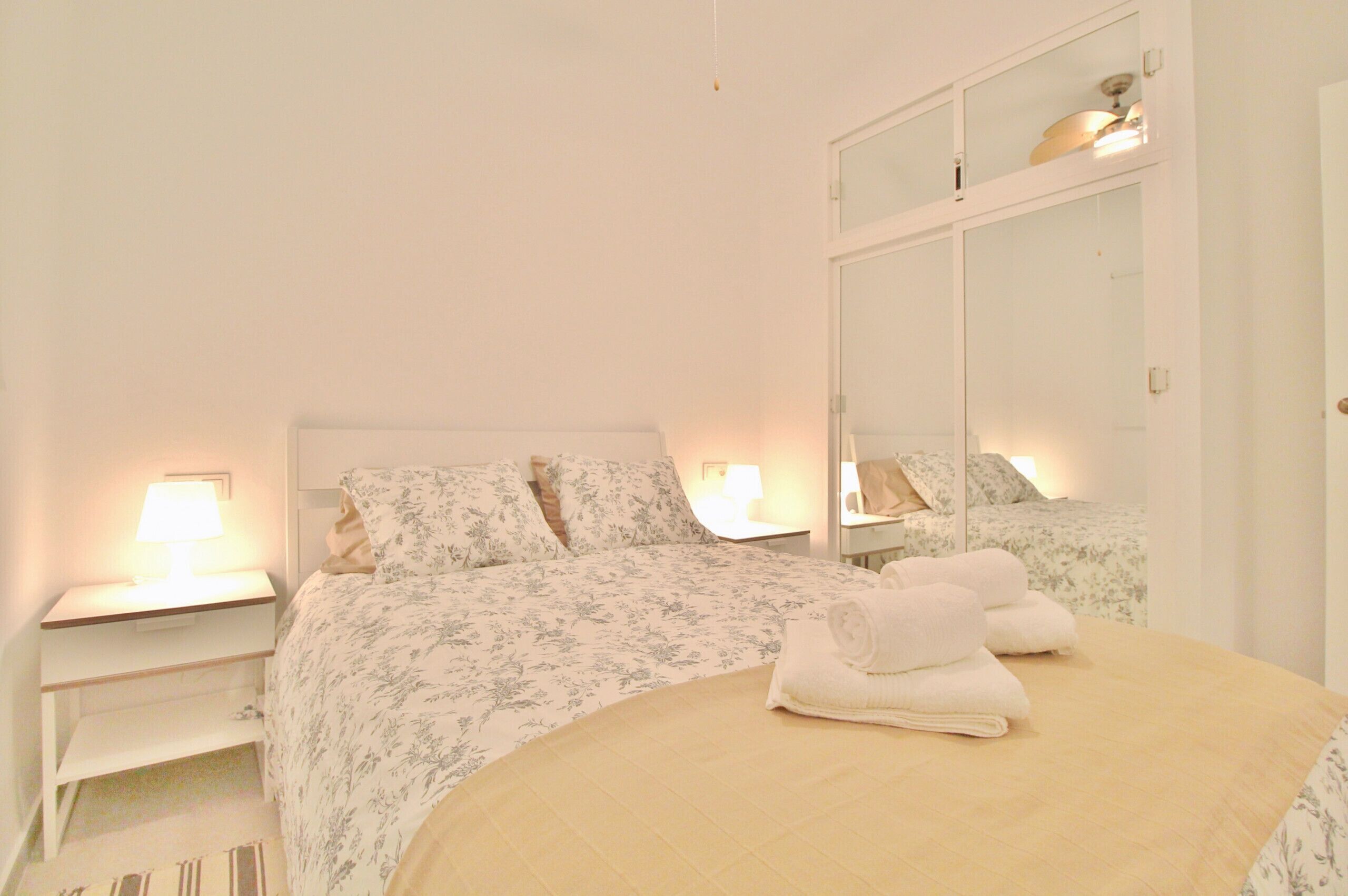 A neatly made bed with floral bedding and a beige comforter, two bedside lamps, a white nightstand, and a large mirror above the bed.