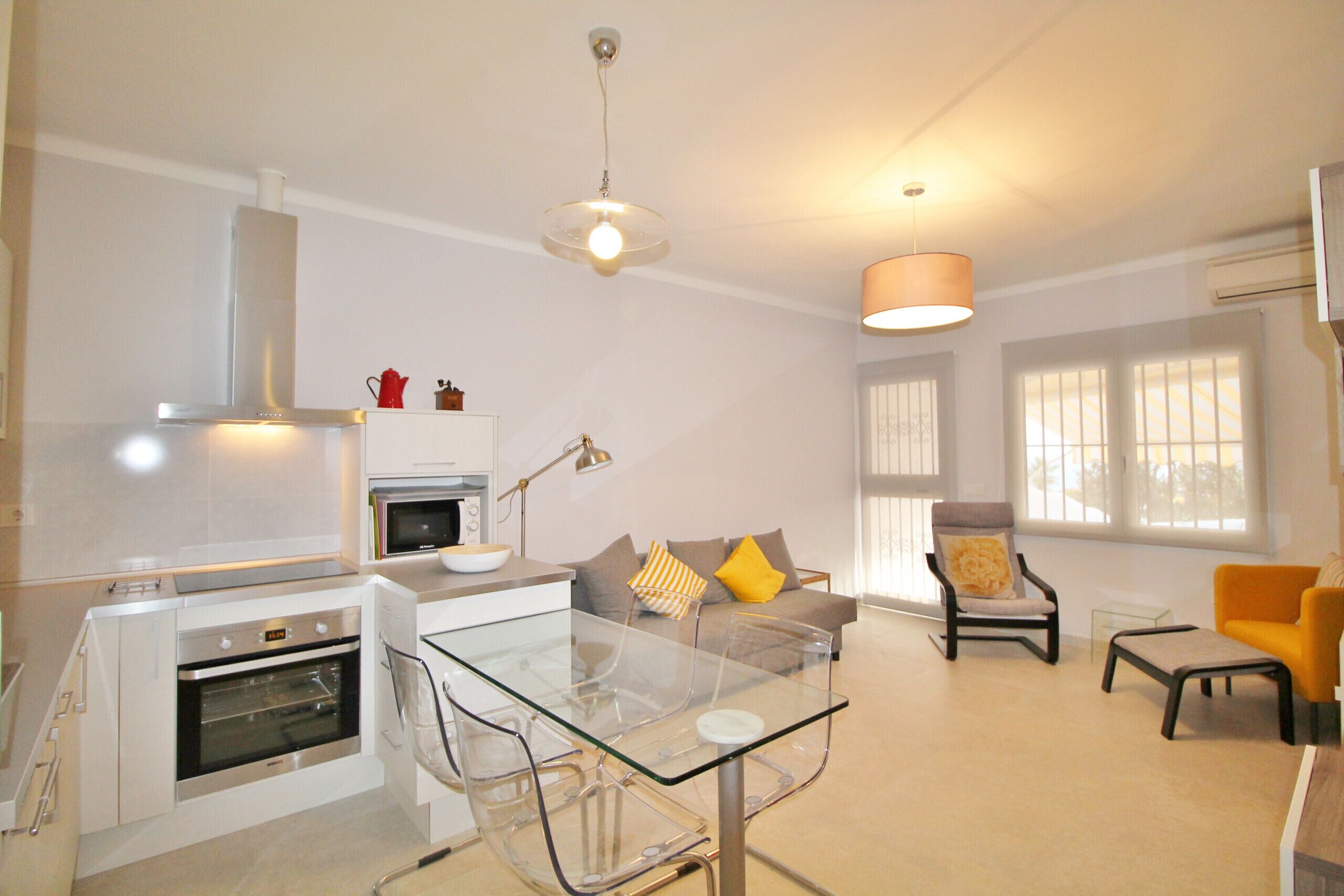 A modern kitchen with a built-in oven and microwave, a glass dining table, and a seating area with a sofa and armchairs.