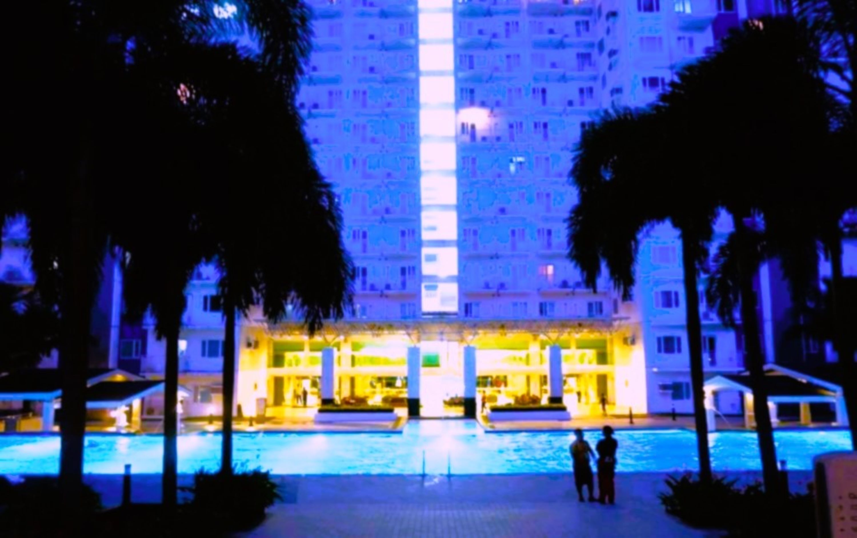 A hotel with a central pool, palm trees, and a brightly lit building facade.