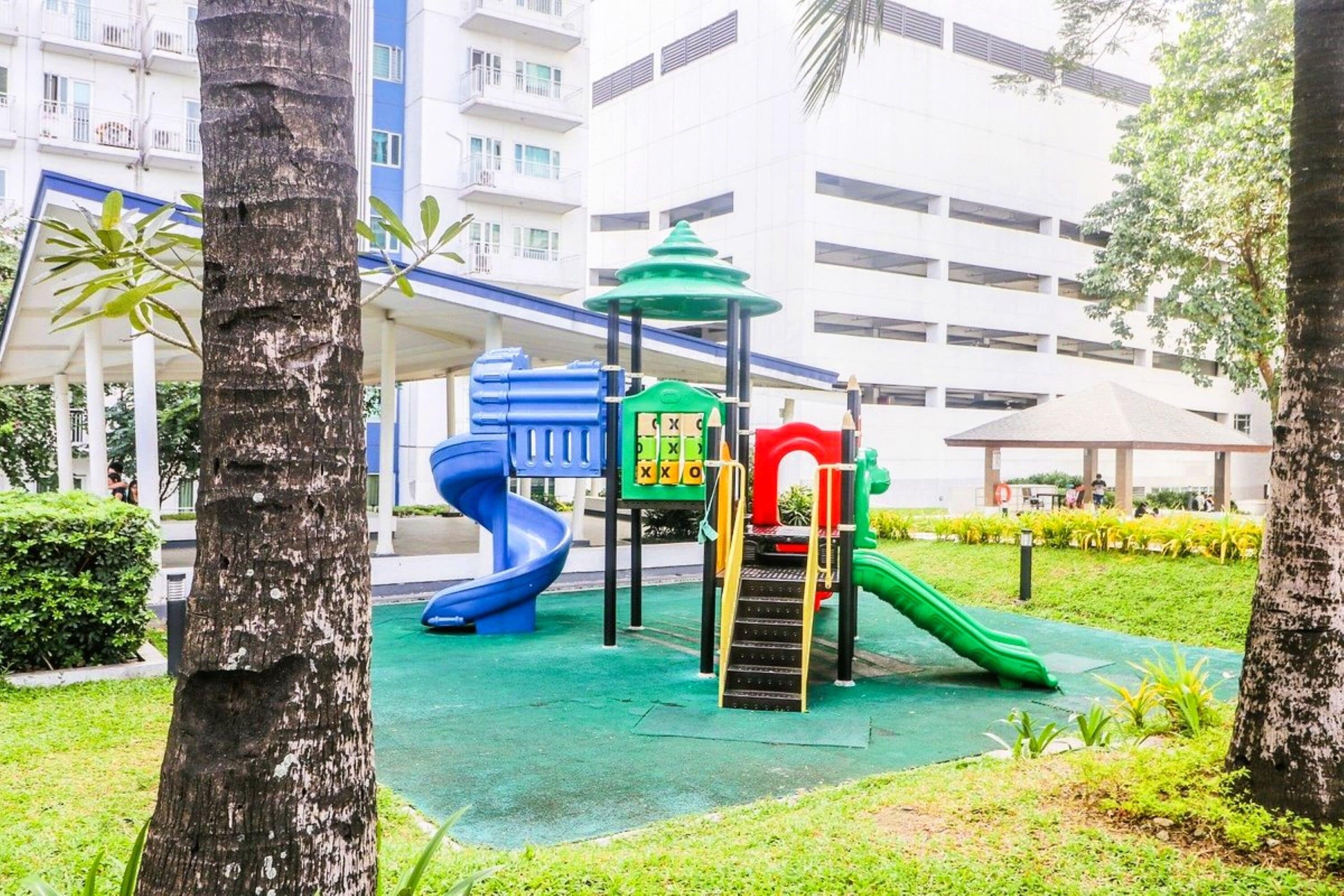 A playground with slides and climbing structures, surrounded by trees and a building in the background.