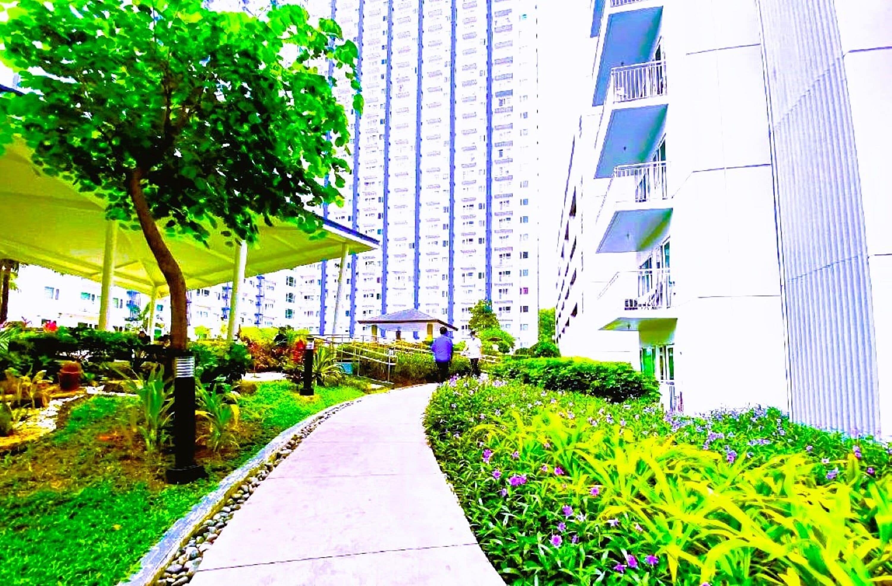 A pathway leading to a residential building with balconies, surrounded by greenery and a covered area.