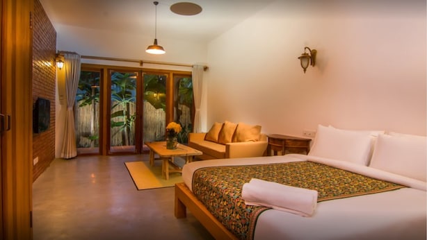 A hotel room with a bed, a sofa, a small table, and a view of tropical plants through the glass doors. - The Orientation Lodge (Siem Reap)