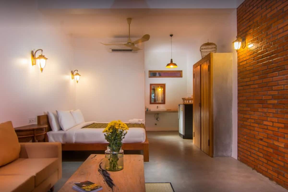 A modern hotel room with a bed, a sofa, a coffee table with a vase of flowers, and a brick wall.
