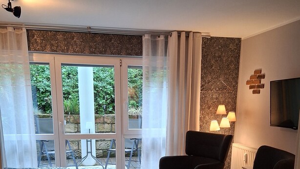 Una sala de estar moderna con un juego de sofá, una mesita y un televisor montado en la pared. Amplias ventanas con cortinas translúcidas permiten el ingreso de luz natural.