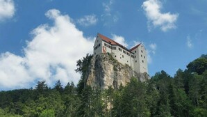 Un castillo enclavado en un acantilado con techo rojo, rodeado de un bosque frondoso.