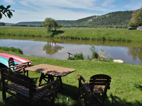 Un montaje para picnic con una mesa y sillas frente a un lago tranquilo, rodeado de exuberante vegetación y colinas al fondo.