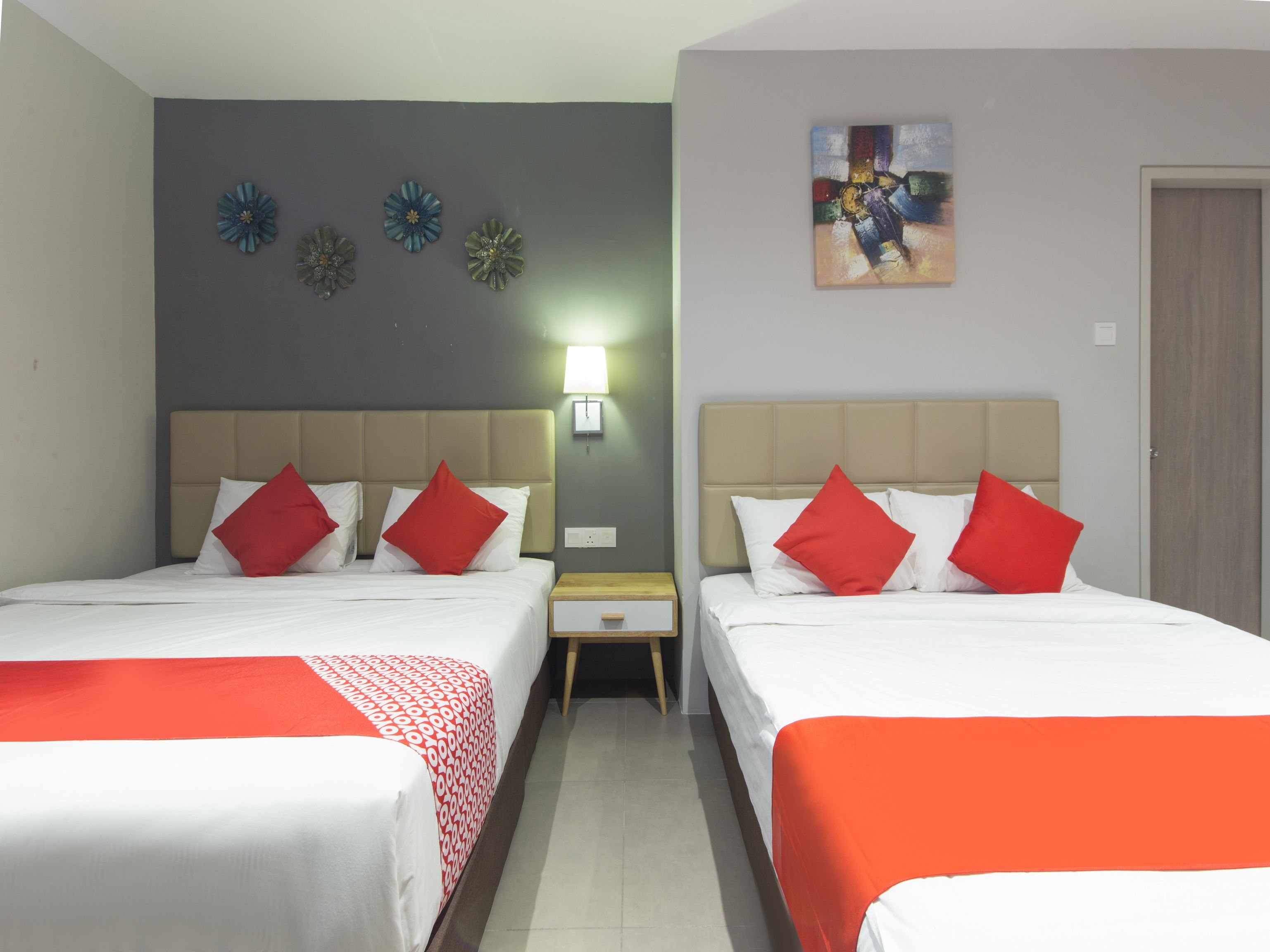 Habitación de hotel con dos camas, cada una con colcha roja y blanca, una mesita de noche con lámpara, y una pared decorada con flores y un cuadro.