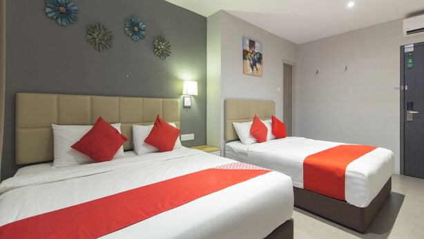 Une chambre d’hôtel avec deux lits, chacun agrémenté de draps blancs et de touches de couleur rouge, un mur gris orné d’un décor floral, un tableau au-dessus des lits et une porte avec une enseigne verte.