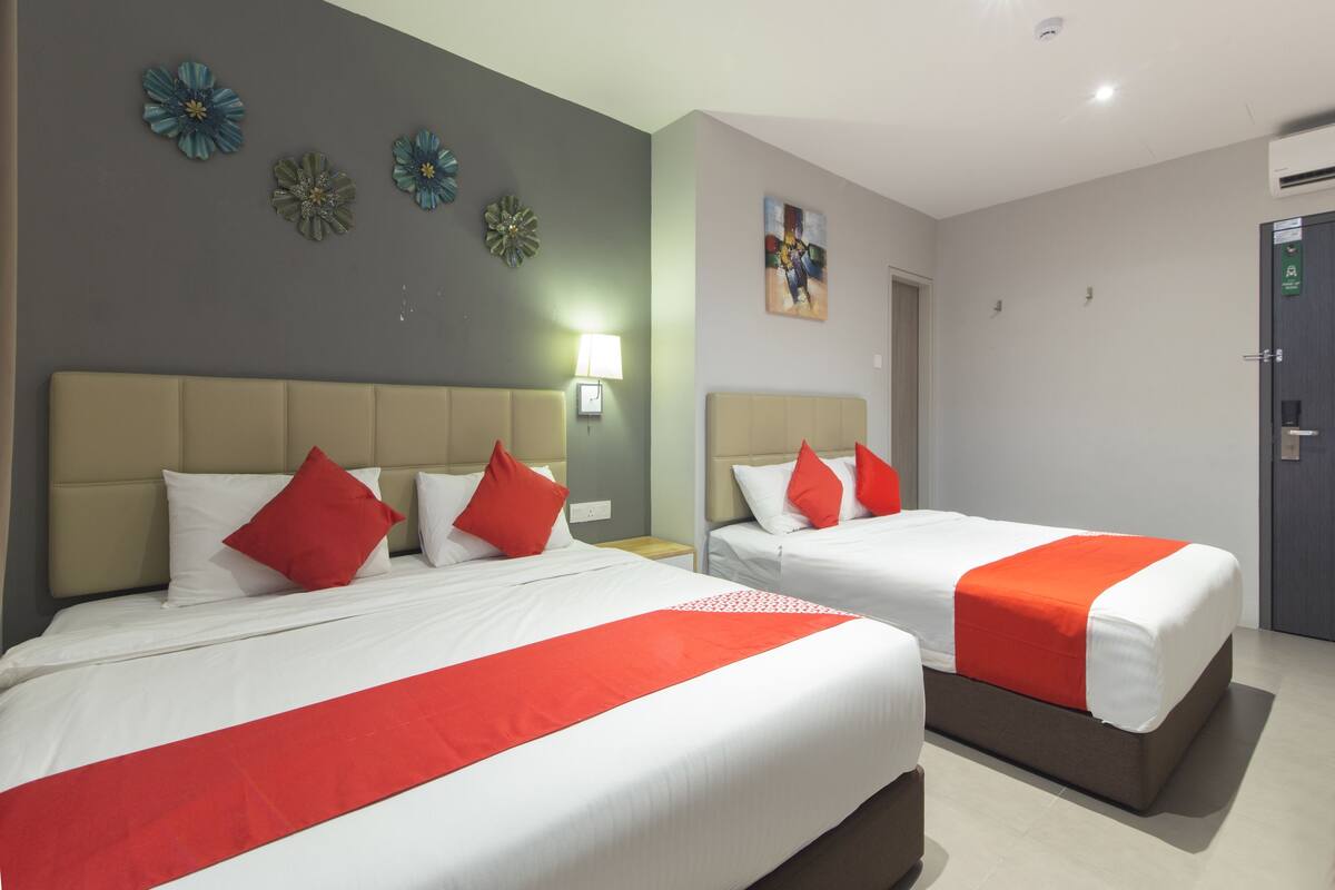 Ein Hotelzimmer mit zwei Betten, jeweils mit weißer Bettwäsche und roten Akzenten, einer grauen Wand mit dekorativer Blumenkunst, einem Gemälde über den Betten und einer Tür mit einem grünen Schild.