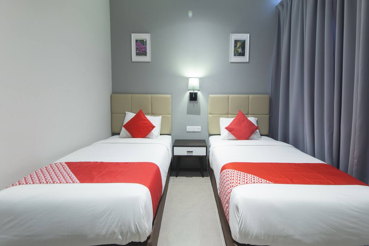 Ein Hotelzimmer mit zwei Betten, jeweils mit einer rot-weißen Bettdecke, einem beigen Kopfteil und einem kleinen Nachttisch mit einer Lampe.