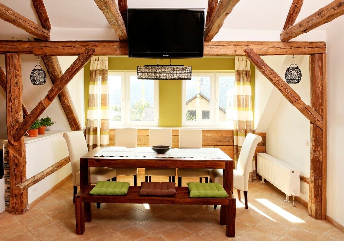 Een eethoek met een houten tafel en banken, een televisie erboven en uitzicht op het exterieur door grote ramen.