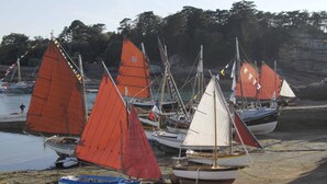 Mehrere Segelboote liegen vor einem Sandstrand an, im Hintergrund sind felsige Klippen zu sehen.
