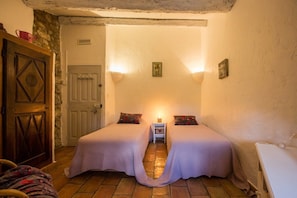 A cozy bedroom with two beds, a stone wall, a wooden door, and a small bedside table with a lamp. - Gîte Lagon I Terrasse I 4 pers. I Domaine le C4 (Berrias-et-Casteljau)