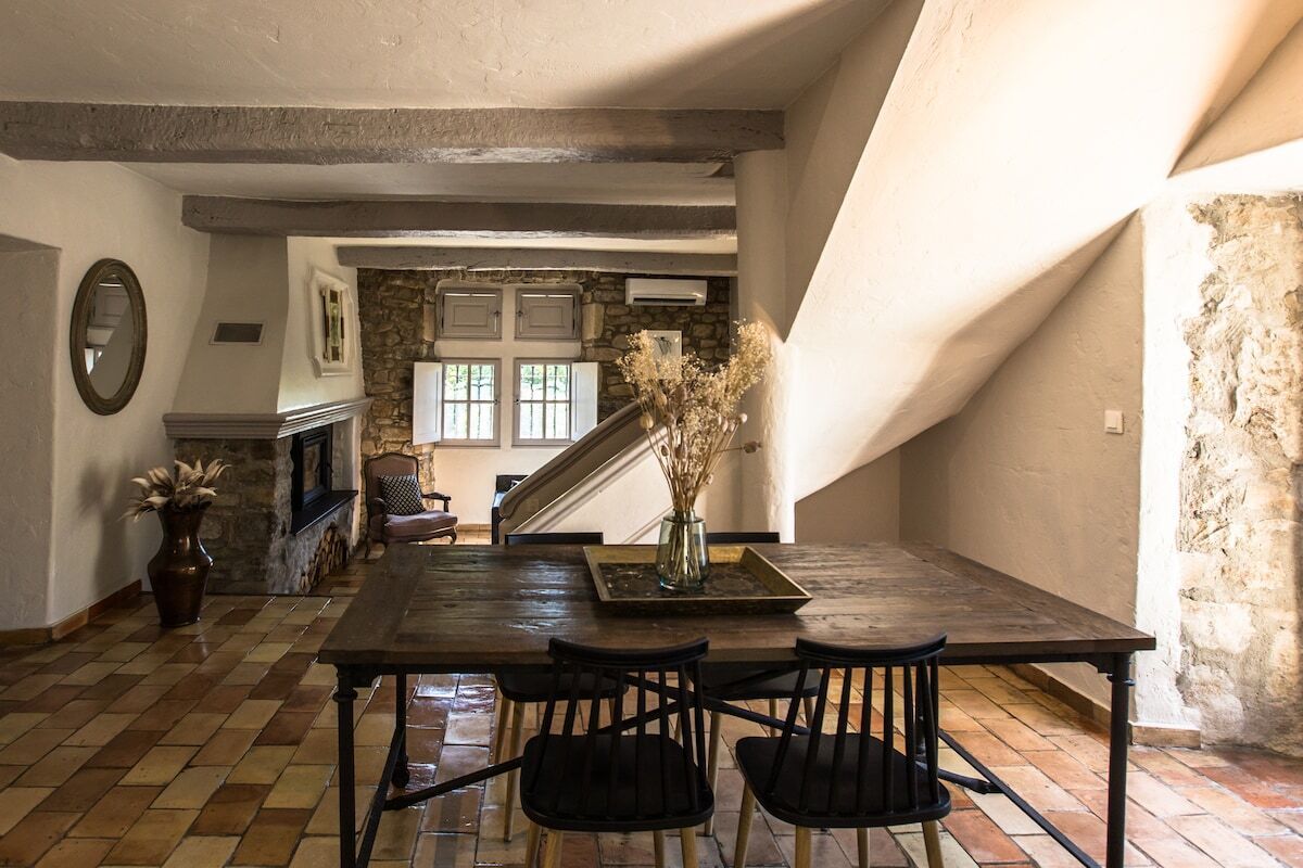 A dining area with a wooden table, black chairs, a vase with dried flowers, and a stone wall in the background.