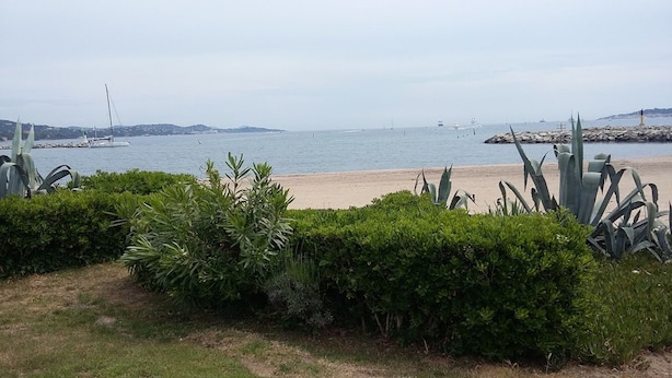 Paisatge costaner amb una platja sorrenca, vegetació verda abundant i una mar tranquil·la amb vaixells.
