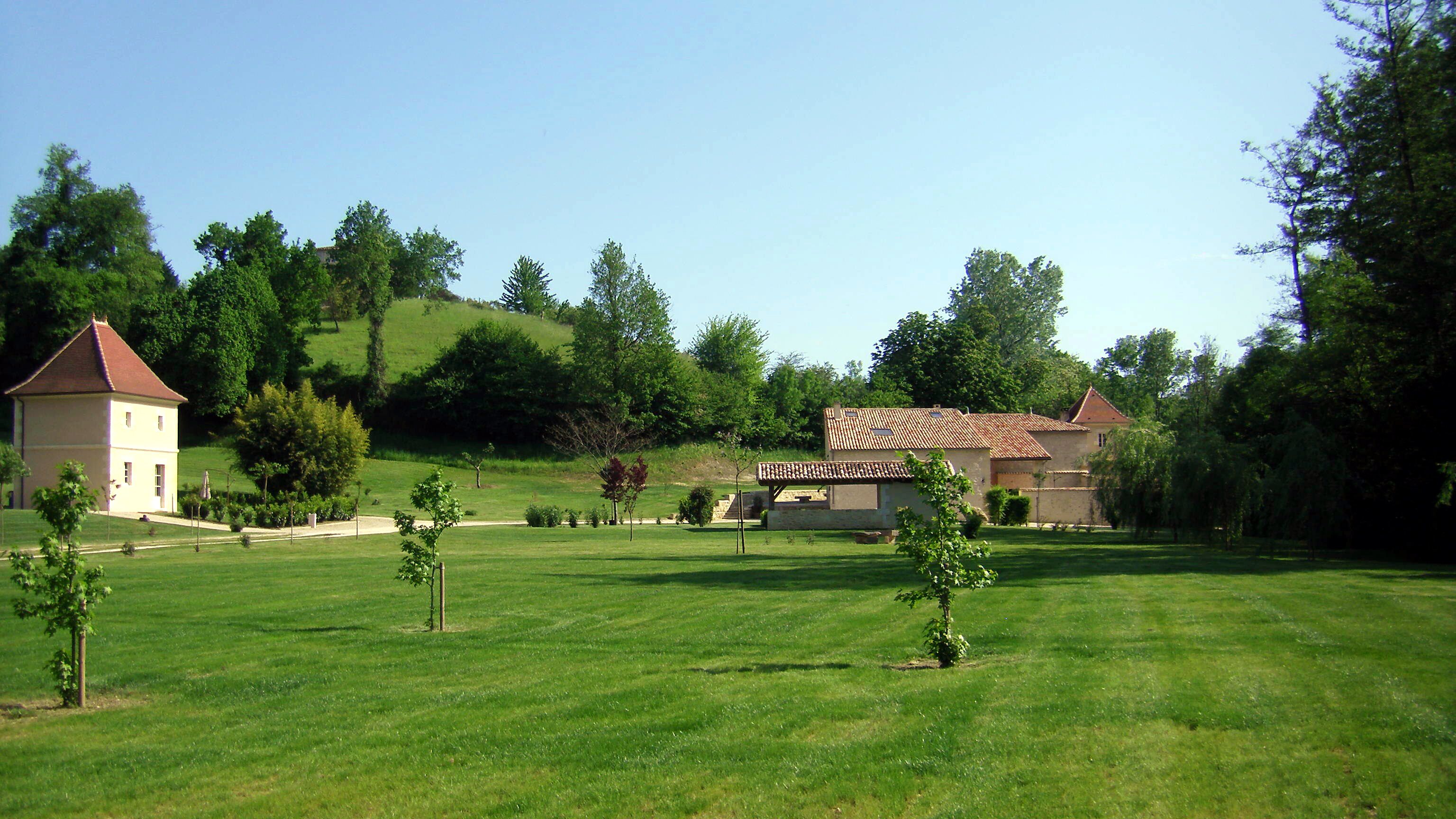 Un paysage rural avec un grand champ verdoyant, deux bâtiments aux toits recouverts de tuiles rouges et des arbres tout autour.