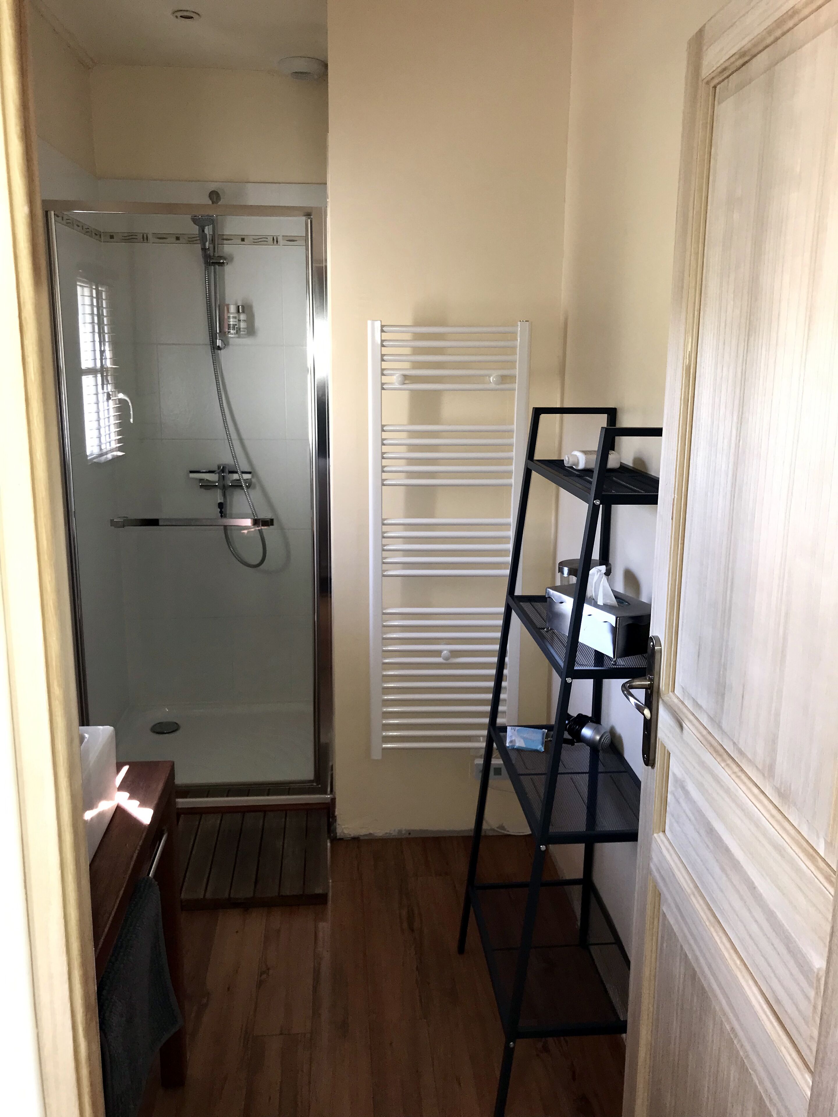 Un baño pequeño con ducha envuelta en vidrio, un toallero blanco y una unidad de estantes de metal negro.