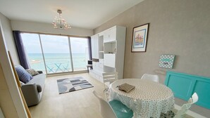Un salon moderne avec un canapé, une table à manger et des chaises, une vue sur la mer grâce à de grandes fenêtres et un luminaire au plafond.
