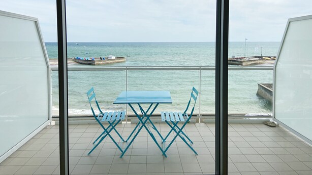 Un balcon avec une table et des chaises, donnant sur une mer calme où l’on peut apercevoir des bateaux et une jetée.