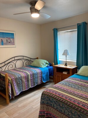A bedroom with two single beds, a ceiling fan, a nightstand with a lamp, a window with curtains, and a painting on the wall.
