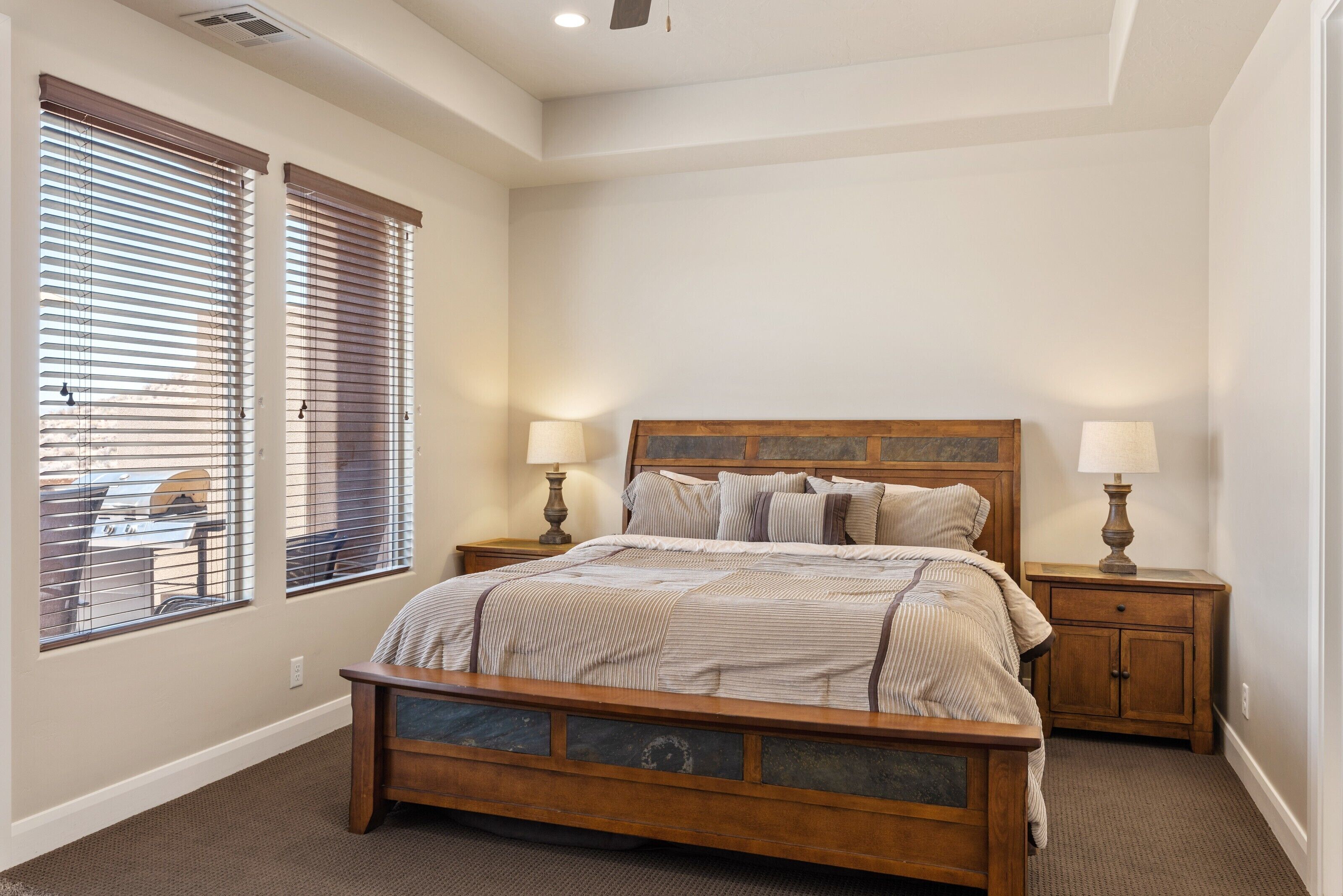 A bedroom with a wooden bed, two bedside tables with lamps, and large windows with blinds.