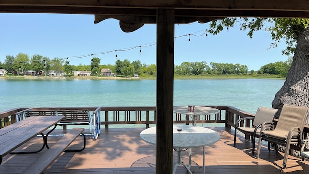 A wooden deck with picnic tables and chairs, overlooking a calm lake with houses in the background. - Gilead Lakefront Cottage with Large Deck and Beautiful View (Bronson)