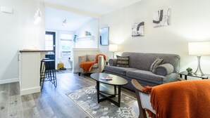 Ein modernes Wohnzimmer mit grauem Sofa, Couchtisch und Barhocker. Im Hintergrund ist eine Küchenzeile mit weißer Arbeitsplatte und Pendelleuchten zu sehen.