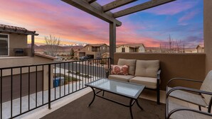 A balcony with a glass table, a sofa, and a chair, overlooking a residential area with houses and mountains in the background. - Desert Pearl at Paradise Village! Waterpark & Lazy River! (Santa Clara)