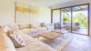 Uma sala de estar moderna com um sofá grande, uma mesa de centro e um ventilador de teto. Há uma porta de vidro deslizante que dá para uma área externa com vista para o verde.