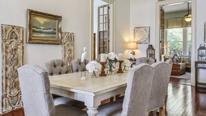 Une salle à manger avec une grande table en bois, six chaises rembourrées, un lustre et une vue sur l’extérieur par une porte.
