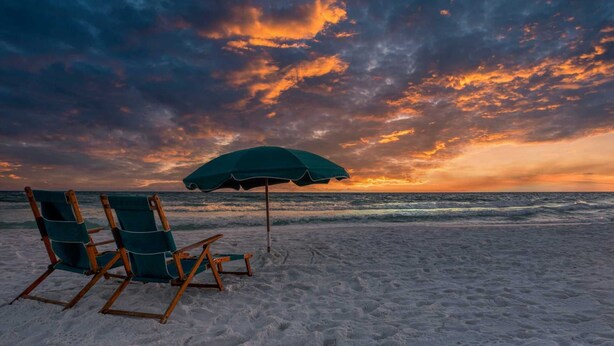 A beach with two wooden lounge chairs and a green umbrella under a sunset sky.