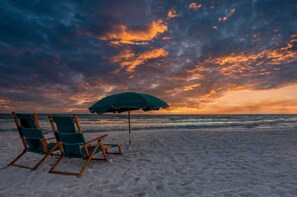 A beach with two wooden lounge chairs and a green umbrella under a sunset sky.