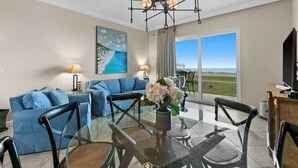 A modern living room with a glass dining table, blue sofa, and a painting of a beach.