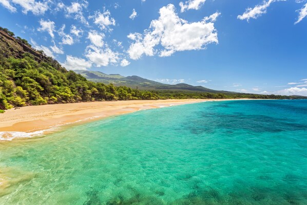 Une plage à l’eau turquoise et limpide, bordée d’un rivage sablonneux, avec des collines verdoyantes en arrière-plan.
